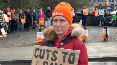Dr Melissa Ryan wearing an orange BMA hat and a red coat stands at a wet roadside holding a cardboard sign reading “Cuts to pay drive doctors away", with other doctors and picket signs behind her.