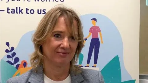 Laura Seebholm is standing in front of a Wearside Women in Need poster and is wearing a light blue blazer with a white T-short underneath.