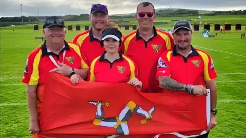 Isle of Man Archery Team Four men and one woman stand together on a green field holding a Manx flag, which is red with a yellow, grey and white triskelion. They wear red T-shirts and four are wearing caps.