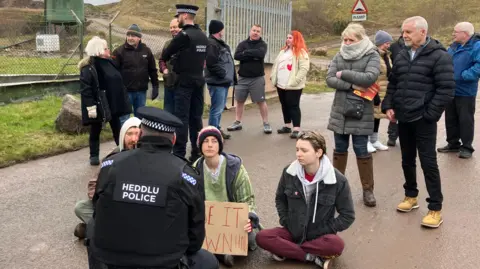 A group of people waiting outside the entrance to the landfill, with police speaking to some protesters that are sat on the ground