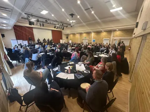 Cad Taylor/BBC Suffolk Large gathering sitting around a number of round tables with black tablecloths looking into the distance at people standing on the stage. Tables have white sheets of paper on them. Stage has red curtains behind and a white background. No faces are distinguishable
