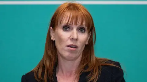 Angela Rayner in a dark suit against a green background