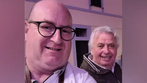 Phil Roberts A selfie showing Dr Phil Roberts and Silvano Casaldi. Phil is at the front left and is wearing black framed glasses and a white shirt. Silvano is behind him and has white hair and is wearing a brown jacket and smiling. 