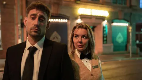 BBC/Element Pictures Left: Matty (played by actor Michael Socha) dressed as a croupier wearing a black suit in TV drama The Cage alongside his co-star Leanne (Sheridan Smith) wearing a mustard coloured waistcoat and white shirt. They are standing outside a casino in Liverpool.