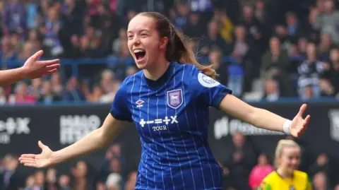 Lucy O'Brien from Ipswich Town, celebrates her goal during the match at Portman Road. She looks away from the camera as she runs. She has brown hair that has been tied back behind her head. She wears a blue Ipswich Town strip.