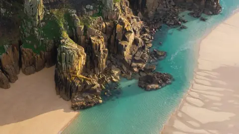 Photo provided by the South West Coast Path Association, Photographer: George Cryer An aerial shot looking down at water running across a sandy beach, next to a rocky terrain. The water is turquoise blue in colour. 