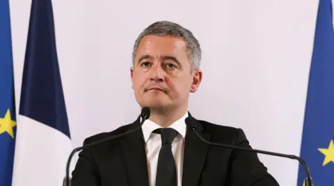 EPA Gérald Dermanin (middle-aged man with short-cropped grey hair) stands in front of two mics on a podium wearing a black suit, white shirt and black tie. Behind him are two EU flags and a French one with the bottom portion out of frame