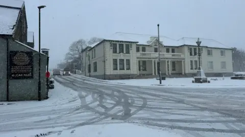 A large building with a balcony in a street covered with snow and tyre marks.