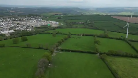 Rolling green fields with a couple of wind turbines in the distance and a new housing development being built 