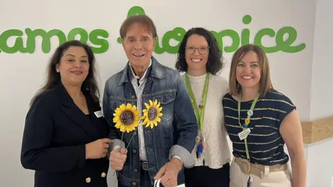 Thames Hospice Sir Cliff (holding two sunflowers and wearing a denim jacket) poses with Sunita Arora, Jane Symmons, and head of marketing Stephanie Peters)