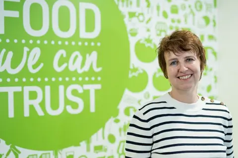 Food Standards Agency Rebecca Sudworth from the Food Standards Agency stood in a black and white stripey jumper in front of a green and white sign about food