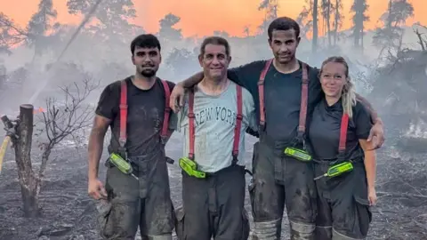 Peterborough Volunteer Fire Brigade Four people are standing in a woodland which is on fire. They are all wearing grey overalls and have a yellow walkie talkie attached to their suspenders. 