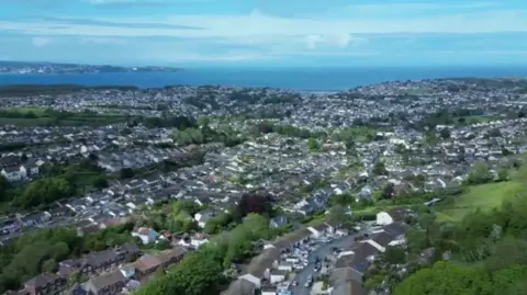 A drone photograph of an area of Brixham which shows lots of houses, roads and some trees as well as the sea further away. 