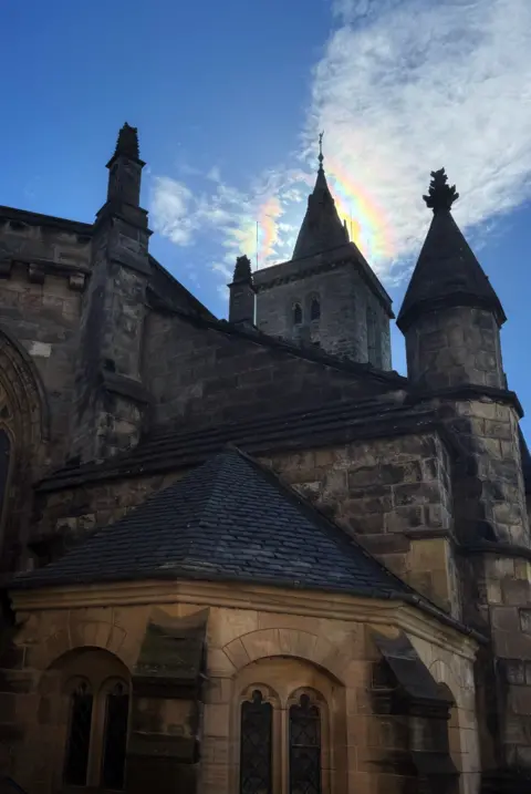 Peter Xu A large chapel with turrets and spires. A rainbow iridescence cloud is above the tower. There are white clouds on the left and the sky is clear blue.