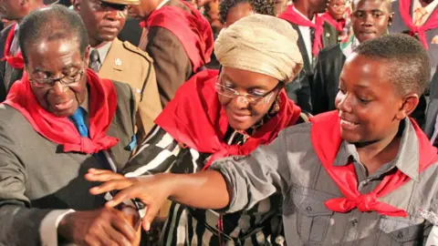 Robert, Grace and Bellarmine Mugabe are standing together, smiling and wearing red neck scarves as they jointly cut a cake in 2011