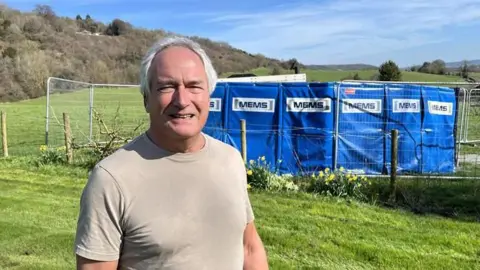 A man with white hair and a beige t-shirt looks at a camera. He is standing in a field, in front of a security fence containing the power generator. It is a sunny day.