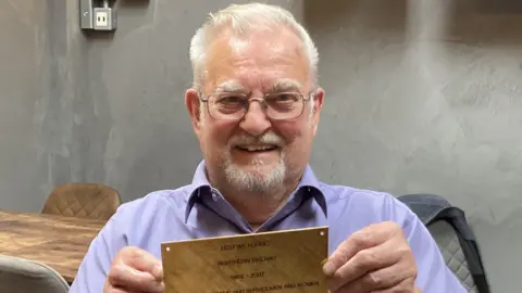 Roger Moseley holding up one of the plaques that will be on the memorial. He is wearing a blue shirt and smiling at the camera