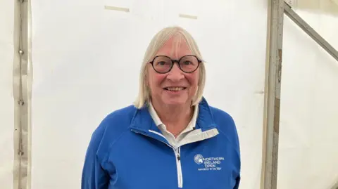 Jackie Neill Tour guide Jackie Neill wearing a blue jumper. She is wearing black glasses