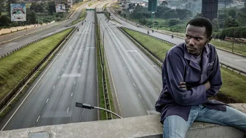 A stranded worker in dark blue top and blue jeans sits on a bridge overlooking an empty Thika highway, one of Kenya's main highways