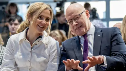 PA Media Mairi McAllan, who has blonde hair, and John Swinney, who is bald with glasses, smile as they speak to each other 