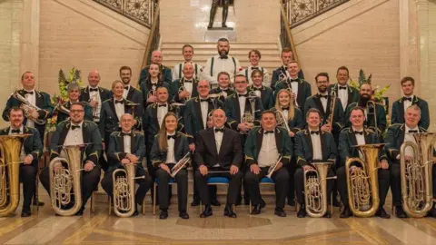 1OB Brass Band The band is sitting and standing on the steps of a marbled hall in Stormont. There is a mixture of ages among the members - men and women between the age of 15 and 72. They are all wearing white shirts, a black bow-tie and black trousers. Most of the members are wearing a green jacket that has gold embroidered embellishments.