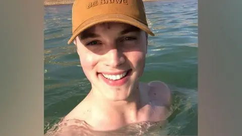 A photo of a man in the sea on a sunny day. There is water up to his shoulders and he is wearing a yellow baseball cap. He is smiling.