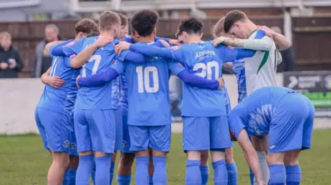Alex Riches Players in light blue strips link arms in a huddle on a pitch with indistinguishable onlookers in the background.