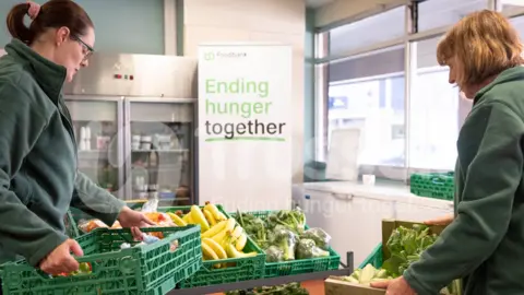 Trussell Two women are holding boxes of fresh food. Bananas, cabbages and broccoli are in boxes.