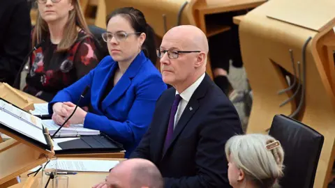 Getty Images The government front bench at Holyrood - with first minister John Swinney in the centre, a bald man with glasses in a dark suit and purple tie. To his left is his deputy, Kate Forbes, a woman with dark hair tied back behind her head, glasses, and a royal blue suit.
