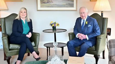 A man and woman sit in green leather chairs, looking at the camera. The woman, who sits on the left, is wearing a green shirt with a navy suit, she wears black high heels. The man wears a blue pinstripe suit with a blue tie and white shirt. He has a yellow flower in his lapel. Behind them is a white wall with three framed paintings. In between them are two small brown, round tables.
In front of them sits a green table with crystal whiskey glasses and jug. 