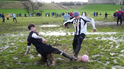 PA Media One man is slide tackling another man in a Newcastle United football kit on a very wet and muddy field. A large ring of spectators can be seen in the distance. A mud spray is coming off the slide-tackling player.