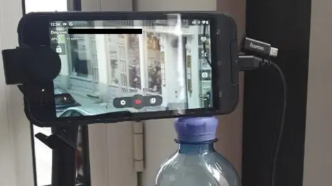 Metropolitan Police handout Propped up with a stand and a water bottle, a phone camera is trained on the front door of Christo Grozev's apartment building in Vienna.