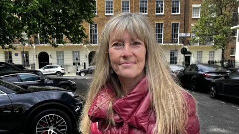 Photo of a woman wearing a red coat and looking directly into the camera. She is standing on a London street.
