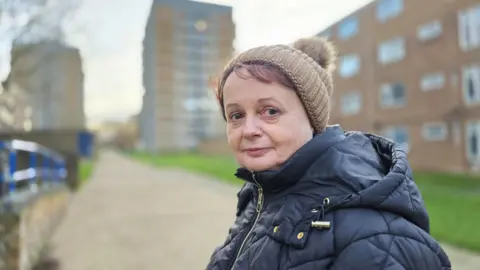 Paul Moseley/BBC Chrissie Rumsby is wearing a dark, padded winter coat and a brown woolly hat. There are a couple of tower blocks, out of focus, in the background.