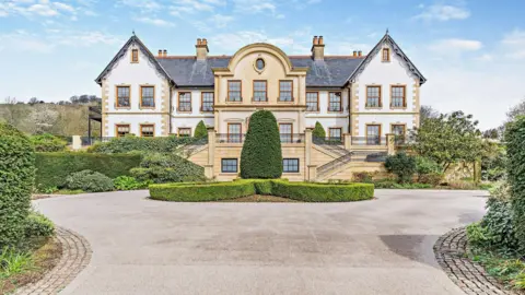 The entrance to a mansion. The property is a beige and white colour with perfectly trimmed bushes in front of the house. A circular driveway can be seen looping around the entrance to the property.