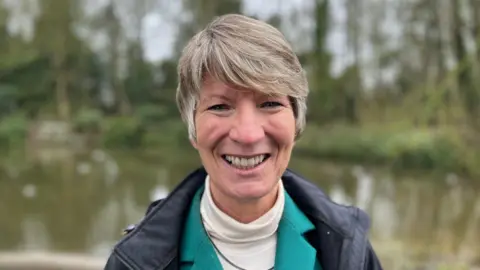 Pippa Heylings has short blonde hair and is wearing a white turtle neck jumper, a green blazer and a navy coat. She is standing in front of a small river which has trees on the other side. 