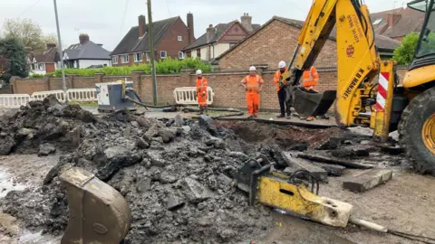 A yellow digger next to a large hole in the road. There is grey rubble around the hole and there are four workers wearing orange high-vis jackets standing on the pavement. There is a residential street with houses in the background.