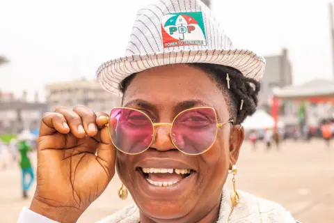 Grace Ekpu / BBC A woman smiles as she holds her right hand up to sunglasses that she is wearing. On her head is a trilby hat with a PDP logo.