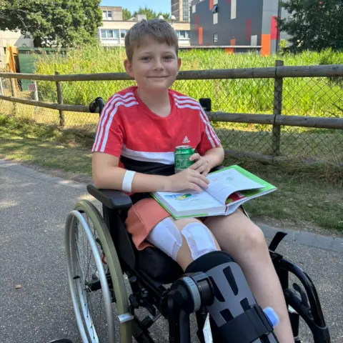 Jo Newman Theo, who has short light brown hair, is wearing a red t-shirt and sat in a wheelchair with a drink can and a book open on his lap. A fence and a patch of long grass can be seen behind him.