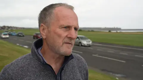 Dave Dixon, campaigner, with short grey hair, wearing a grey fleece, looks to the right as he stands on a grass field, with a road with cars driving past next to him. 