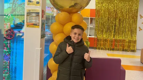 Children with Cancer UK A young boy in a black puffer coat is smiling at the camera with his thumbs up. He is standing in front of a cluster of yellow and gold balloons