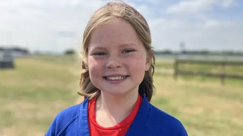 Alex Dunlop/BBC Emily is standing outside and is looking directly at the camera and smiling. She is wearing a red T-shirt and blue cardigan.