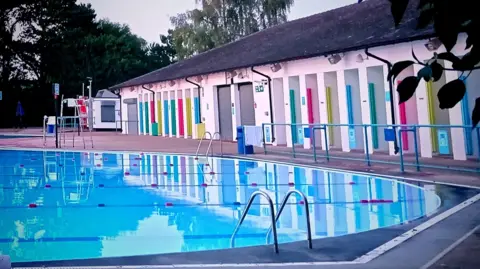 Elsie from the block An outdoor swimming pool with lanes can be seen in the foreground. A white building housing the changing booths cuts through the image with multi-coloured doors with a walkway in front of it before the water's edge.