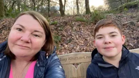 Nicky Wake A woman, Nicky, wears a pink t-shirt and rain coat. Her son, Finn, smiles in a black waterproof. In the selfie they are sitting on a bench in a wooded area. There are orange leaves on the floor and it appears to be autumn.