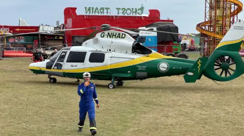 BBC An air ambulance is landed on a grassed area in front of several fairground rides. A member of the emergency services can be seen in the foreground. He is wearing a blue uniform and white helmet.