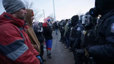 Demonstrators stand in a row in front of members of US Customs and Border Protection and other law enforcement officials, near the Bishop Henry Whipple Federal Building. Federal agents are dressed in black and wearing gas masks that cover their face. Demonstrators are wearing plain clothes. Some in the back are waving an upside-down American flag.
