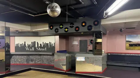 Chris Morgan A mirror panelled DJ booth, with vinyl records stuck on the wall, stands next to an image of the New York skyline with the words 'Wall Street' on, the final name of the club before it closed