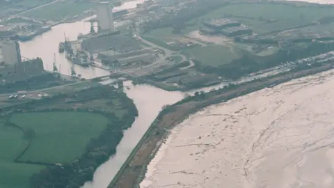 PA A grainy aerial shot of Sharpness Docks, a small dockyard along the Severn Estuary, from 1993. There are fields surrounding the docks and a bridge over the canal, which is separated from the tidal river by a strip of land.