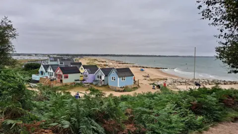 A beach scene framed by green heather. The beach has a row of colourful beach huts, the closest is blue and behind they are purple, green and yellow. There are two small boats on the beach. To the right the sea is blue with some white waves. And to the left you can see sailing boats moored in Christchurch harbour.