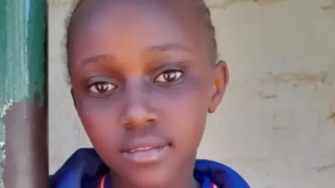 Bridgit Njoki stares at a camera, wearing a top with a blue collar. She stands in front of a white wall.
